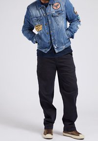 Muž oblečený v modré džínové bundě s nášivkami, černých kalhotách a hnědých teniskách stojí s rukama v kapsách na jednobarevném pozadí.