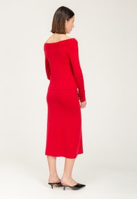 Vestido rojo de punto, con escote off-the-shoulder, mangas largas, cuerpo ajustado y falda a la altura de las rodillas. Zapatos negros sin cordones con puntera puntiaguda.
