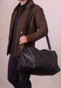 Black leather duffel bag with a textured finish, double zipper closure, and adjustable fabric strap. Model holds it with one hand.