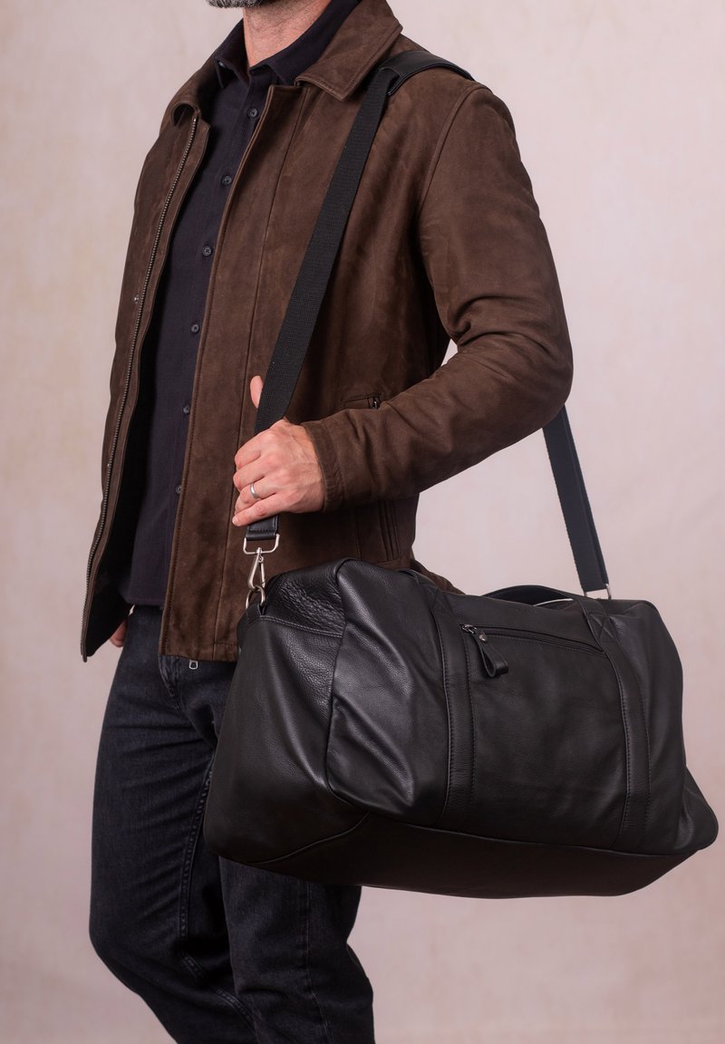 Black leather duffel bag with a textured finish, double zipper closure, and adjustable fabric strap. Model holds it with one hand.