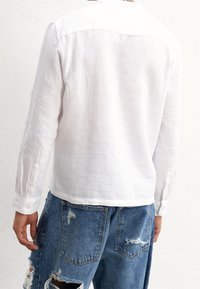 Camicia bianca in lino a maniche lunghe con collo, texture liscia e polsini abbottonati, abbinata a jeans in denim blu slavato con grandi tasche.