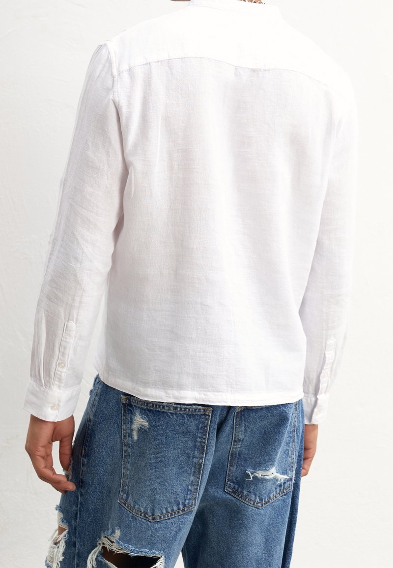 Camicia bianca in lino a maniche lunghe con collo, texture liscia e polsini abbottonati, abbinata a jeans in denim blu slavato con grandi tasche.