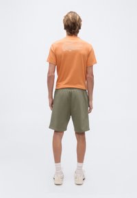 Νέος/Νέα στέκεται, φοράει πορτοκαλί μπλουζάκι με λευκό λογότυπο κροκόδειλου στην πλάτη, χακί σορτς, λευκές κάλτσες και μπεζ αθλητικά παπούτσια.