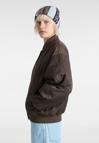 Braune gesteppten Bomberjacke mit gerippten Bündchen, ausgestattet mit Seitentaschen und glatter Textur, kombiniert mit einer mehrfarbigen Strickmütze.