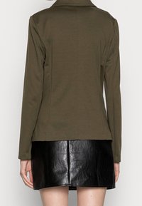 Olivově zelené saka na míru s dlouhými rukávy a klopami s výstřihem, spárované s lesklou černou koženou minisukní, která zdůrazňuje kontrast textur.