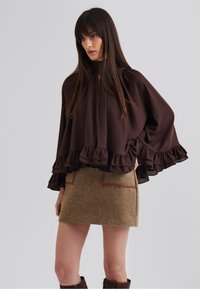 Blouse marron à volants avec un col haut, associée à une jupe beige texturée avec des accents marron et deux poches avant.
