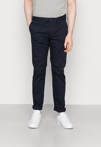 Navyblaue Chinos aus Baumwolle mit geradem Bein, ausgestattet mit einem Knopfverschluss und umgeschlagenen Bündchen, kombiniert mit weißen Sneakers.
