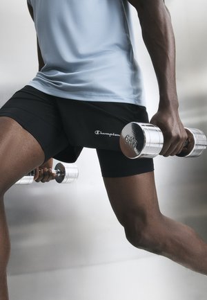 Persona che indossa pantaloncini neri e maglietta azzurra tiene manubri da 6 kg in entrambe le mani, eseguendo un esercizio di affondo o squat diviso.