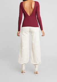 Top bordeaux a maniche lunghe con scollatura a V profonda sul retro, abbinato a pantaloni cream a gamba larga. L'outfit presenta texture e forme fitted in vita.
