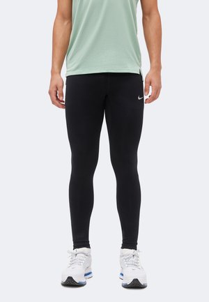 Άτομο με μαύρα κολάν Nike, ανοιχτό πράσινο αμάνικο τοπ και λευκά αθλητικά Nike, που στέκεται μπροστά σε απλό ανοιχτό φόντο.