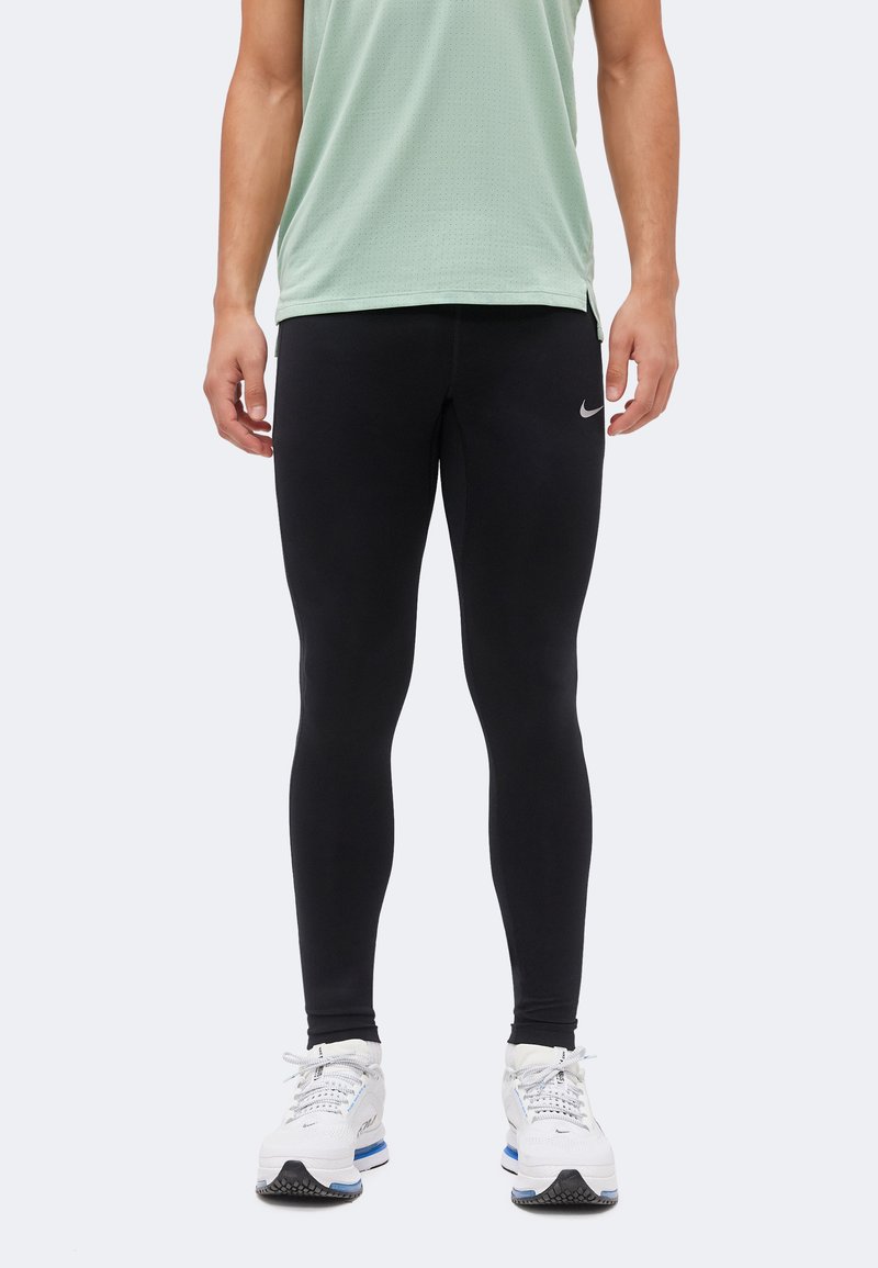 Άτομο με μαύρα κολάν Nike, ανοιχτό πράσινο αμάνικο τοπ και λευκά αθλητικά Nike, που στέκεται μπροστά σε απλό ανοιχτό φόντο.