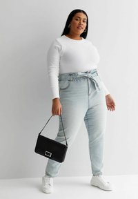 Top blanco de manga larga, vaqueros de tiro alto en azul claro con un cinturón de lazo, zapatillas blancas y un bolso negro. Textura suave, diseño casual.