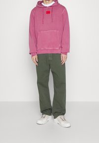 Różowa bluza z kapturem, z kieszenią z przodu i czerwoną naszywką logo, w połączeniu z zielonymi spodniami i białymi trampkami. Styl casualowy i luźny krój.