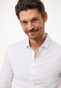 Camicia bianca con colletto standard, tessuto liscio e design aderente, dotata di due bottoni al collo e maniche lunghe.