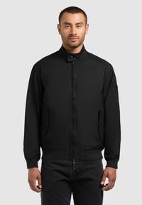 Lühikeste juustega mees, kes kannab musta lukuga jopega, millel on püstitatud krae ja nööbitavad taskukatted, seisab ühetaolise heleda tausta ees.