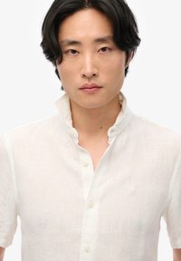 Biała lniana koszula z krótkim rękawem i stójką, z zapięciem na guziki oraz subtelną fakturą; małe haftowane logo na lewej piersi.