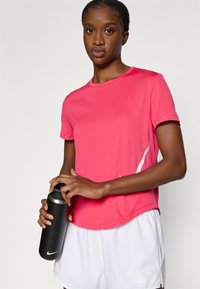 Ροζ αθλητική μπλούζα με στρογγυλή λαιμόκοψη, κοντά μανίκια και λευκή λεπτομέρεια. Κρατάει μαύρο αθλητικό παγούρι με ασημένιο καπάκι.