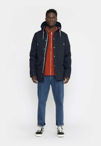 Navy blue jacket with a hood, silver snap buttons, two front pockets, layered over an orange shirt and paired with blue jeans and black sneakers.