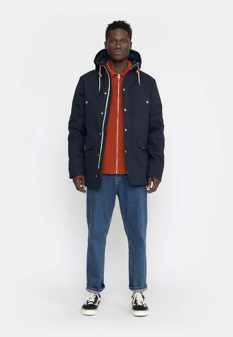Navy blue jacket with a hood, silver snap buttons, two front pockets, layered over an orange shirt and paired with blue jeans and black sneakers.
