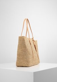 Borsa a spalla intrecciata in paglia con manici in pelle liscia color cuoio. Forma rettangolare, colore beige naturale, con un dettaglio annodato su un lato.
