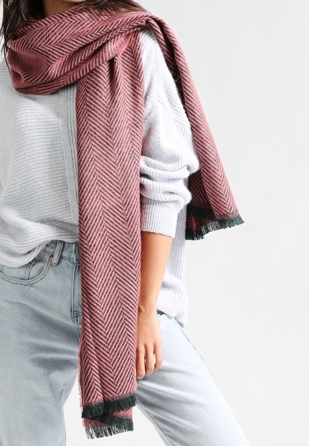 Femme portant un pull gris clair et un jean bleu clair, avec une longue écharpe rose à chevrons drapée sur l'épaule.