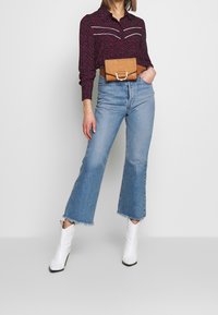 Camicia scura a fantasia con accenti bianchi, jeans svasati a vita alta, pochette in pelle marrone e stivaletti bianchi con punta a punta. Materiali testurizzati visibili.