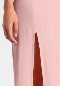 Robe rose clair en tissu texturé, présentant une fente haute sur un côté et une silhouette ajustée qui met en valeur l'ensemble du design.