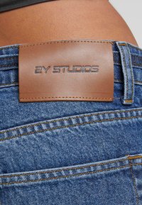 Kratki jedan kadrovni prikaz stražnjeg džepa traperica s ušivenom smeđom kožnom etiketom na kojoj piše "2Y STUDIOS" na pozadini golih poteza kože.