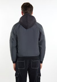 Jachetă bomber gri cu glugă neagră, mansete și tiv cu nervuri, având un finisaj texturat și detalii cu patch-uri pe umeri. Purtată deasupra unor pantaloni închiși la culoare.