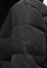 Μανίκι μαύρου καπιτονέ μπουφάν με μεταλλικό έμβλημα "RL" ραμμένο στο πάνω μέρος του μπράτσου.