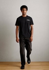 Schwarzes Baumwoll-T-Shirt mit kurzen Ärmeln und Logo auf der Brust, kombiniert mit dunklen Jeans und schwarzen Schuhen, stehend auf einem Holzboden vor einem grauen Hintergrund.