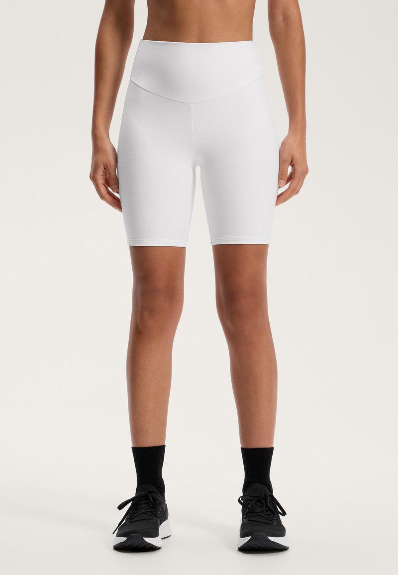 Pantaloni scurți albi, cu talie înaltă, dintr-un material neted, cu un design ajustat și lungime până la mijlocul coapsei, asortați cu încălțăminte sport neagră și șosete.