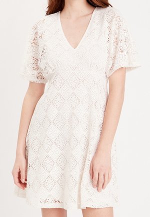 Vrouw draagt een witte kanten jurk met korte pofmouwen en een V-hals, met ingewikkelde bloemen- en geometrische patronen.