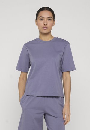 Femme aux cheveux foncés attachés, portant un t-shirt violet uni à manches courtes et un short assorti, debout avec une main dans la poche.