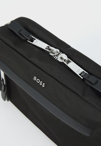 Čierna textúrovaná látková taška so striebornými doplnkami, ktorá má výrazný zips a elegantný dizajn, označená značkou "BOSS" na prednej strane.