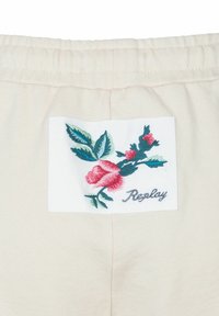 Crème sweatshirtstof met een geborduurd patch van een roze roos en groene bladeren, samen met "Replay" in cursieve letters.