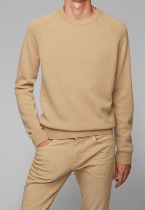 Pull en tricot beige avec un col rond, des poignets et un ourlet côtelés, présentant un motif texturé et une coupe ajustée. Assortie à un pantalon beige coordonné.