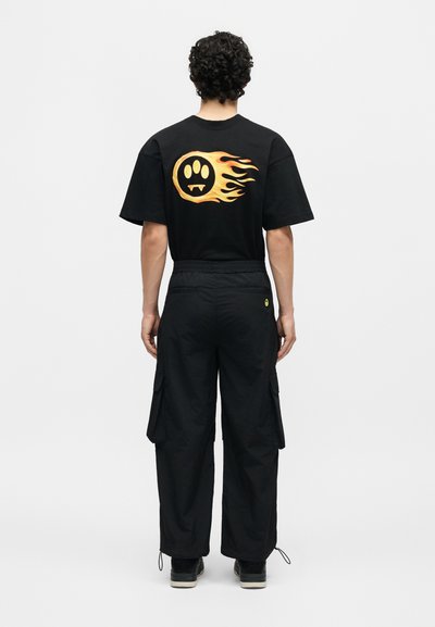 Άτομο με σγουρά μαλλιά που φοράει μαύρο παντελόνι καργκό και μαύρο T-shirt με φλεγόμενο κίτρινο αποτύπωμα πατούσας με κυνόδοντες στην πλάτη.