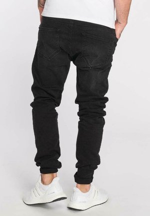 Homme portant un pantalon de jogging noir et des baskets blanches Adidas Ultra Boost, debout les mains dans les poches, de dos.