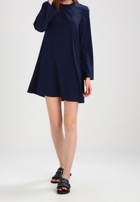 Marinblå A-linjeformad klänning med långa ärmar, tillverkad av ett mjukt tyg. Kombinerad med svarta sandaler med glidfunktion som har dekorativa detaljer.