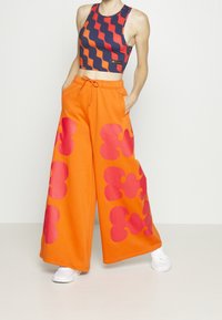 Donna che indossa un crop top senza maniche con strisce ondulate arancioni e nere e pantaloni arancioni larghi con motivi astratti rosa, mani nelle tasche.