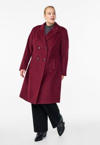 Burgundský vlněný kabát s dvojitým zapínáním, velkými knoflíky, klopovým límcem, bočními kapsami a rovný střih; nošený přes pruhovaný top a černé kalhoty.
