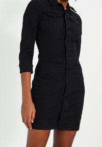 Kobieta ubrana w dopasowaną czarną sukienkę koszulową z guzikami, o rękawach do łokcia, z kieszeniami na piersi i z zapięciem na guziki z przodu.