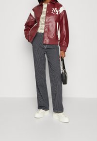 Veste varsity en cuir marron avec accents blancs et logo NY, associée à un pantalon large à rayures et des baskets blanches à plateforme, tenant un sac noir.