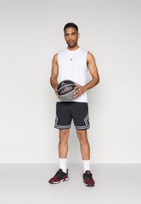 Joueur de basketball portant un maillot sans manches blanc, un short noir avec des accents blancs, et des chaussures de basketball noires et rouges, tenant un ballon de basketball.