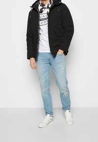 Svart puffjacka med huva, med framfickor och silverdragkedja. Bärs över en vit t-shirt med grafik och ljusblå rivna jeans.