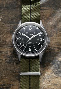 Timex MK1 Automatisch horloge met zwarte wijzerplaat, witte cijfers, zilverkleurige wijzers en groene stoffen band, rustend op een houten oppervlak.