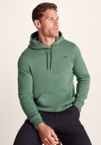 Green hoodie with a front pocket, drawstring hood, and a small brand label on the chest. Worn with dark pants, smooth texture.