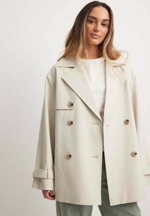Beige kahepoolne jakk, mis on valmistatud siledast kangast, millel on suured nööbid, laiad kraed ja lõdvad varrukad kinnitusribadega.