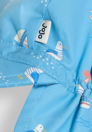 Casquette en tissu bleu clair avec un motif nautique de poissons et de bateaux, avec un élastique à l'arrière et une petite étiquette cousue sur le côté.